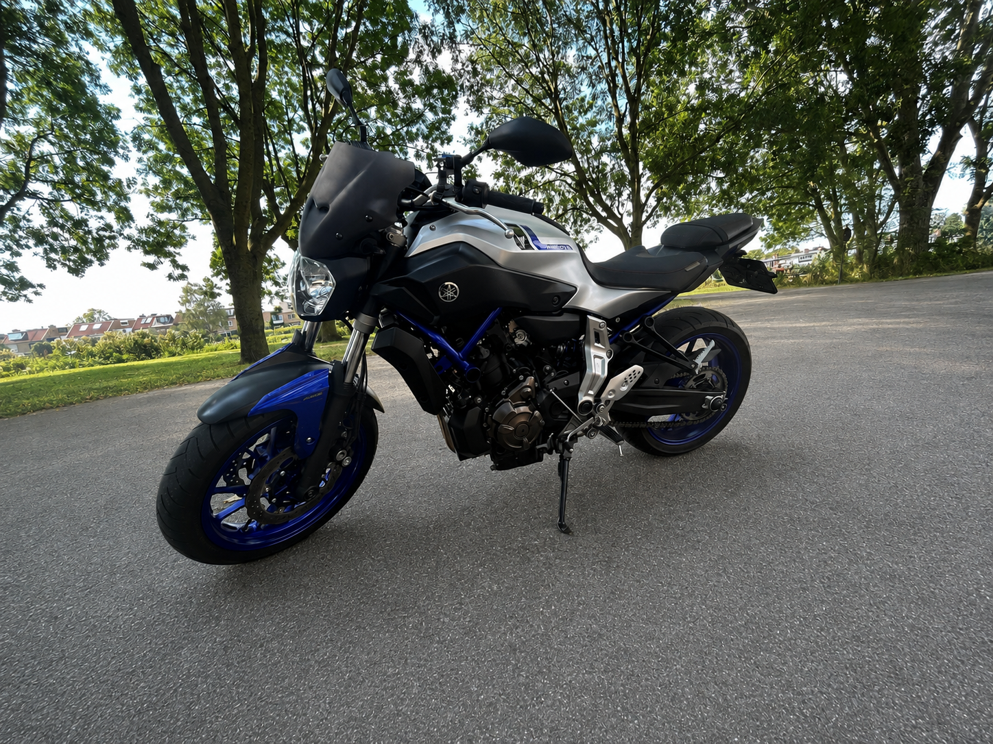 Een zwarte en zilveren motorfiets met blauwe velgen geparkeerd op een asfaltweg onder groene bomen.