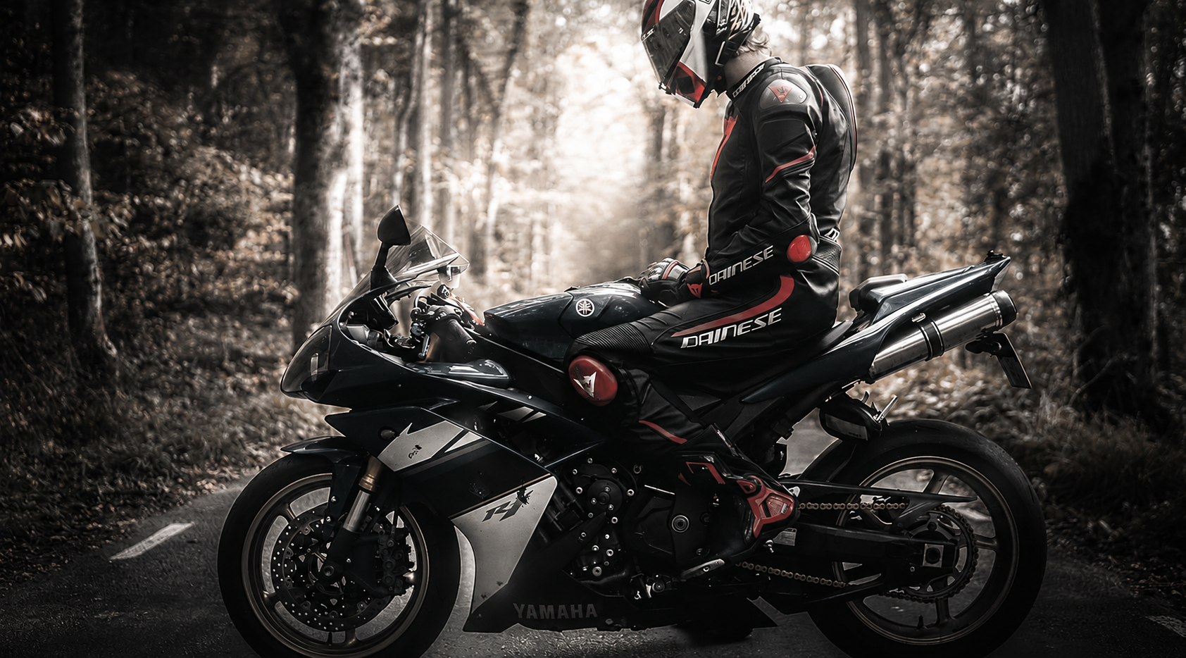 Een persoon in beschermende motoroutfit en helm zit op een zwarte Yamaha sportmotor op een bosweg, omgeven door bomen.