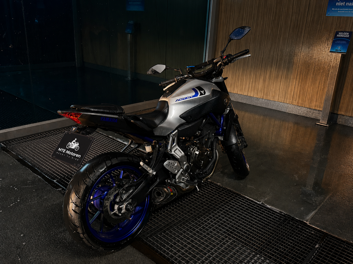 Zwarte en zilveren Yamaha motorfiets met blauwe accenten, gepositioneerd op een natte vloer binnen een modern gebouw, met een bord dat zegt 'MTE Motoren'.