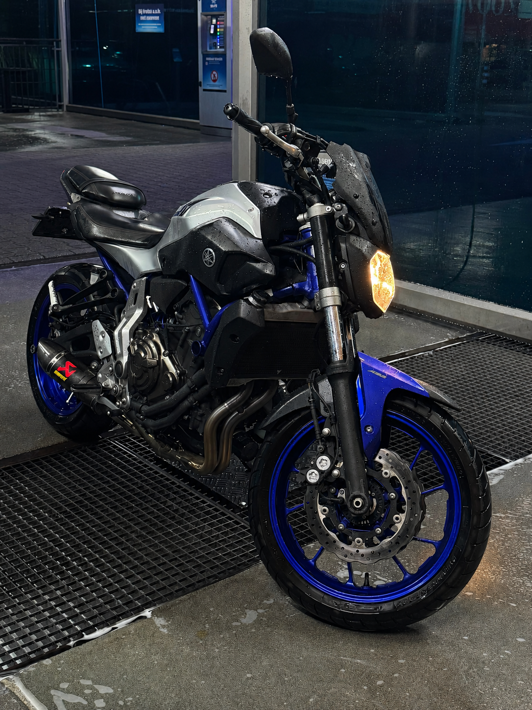 Mooie Yamaha motorfiets in het zwart en blauw, nat door regen, geparkeerd bij een oplaadpunt of wachtruimte buiten bij een autodepot of tankstation, met een regenbouwwind en een stedelijke achtergrond.