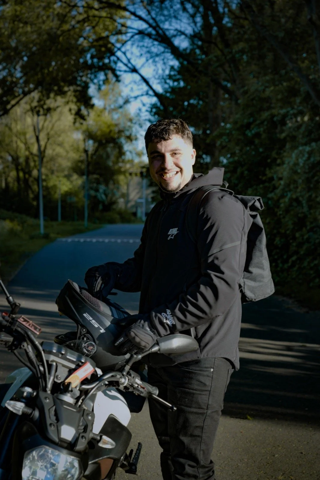 Lachende man in zwart jack en handschoenen staat naast een motorfiets in een bosrijke omgeving met bomen en een weg.