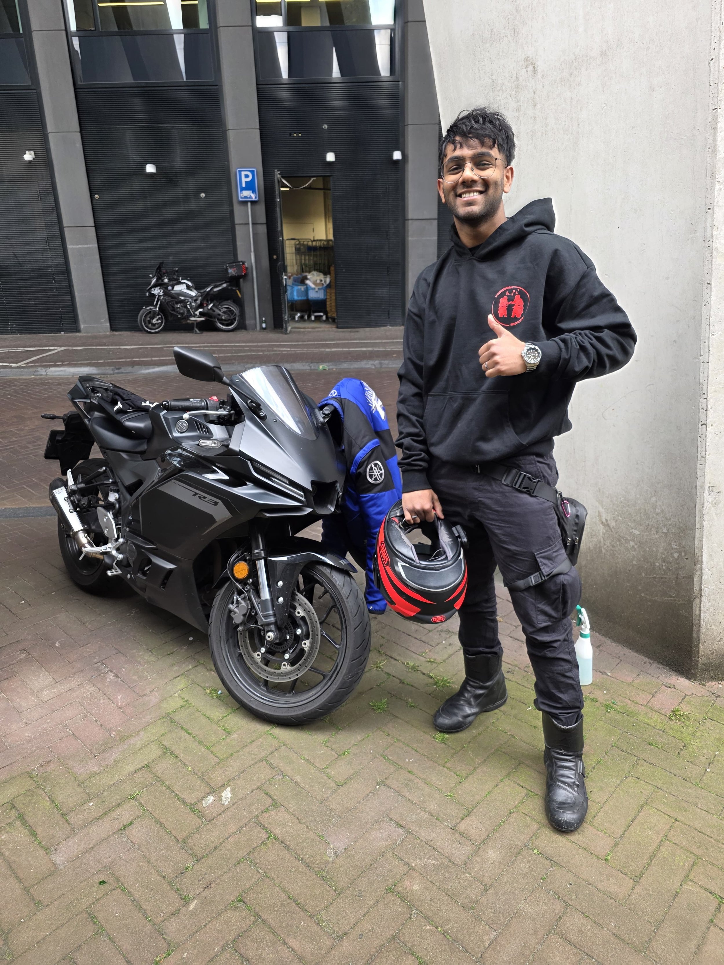 Vrolijke man met bril en masker in hand naast een sportmotor, staand tegen een muur op straat, in de achtergrond een gebouw met parkings.