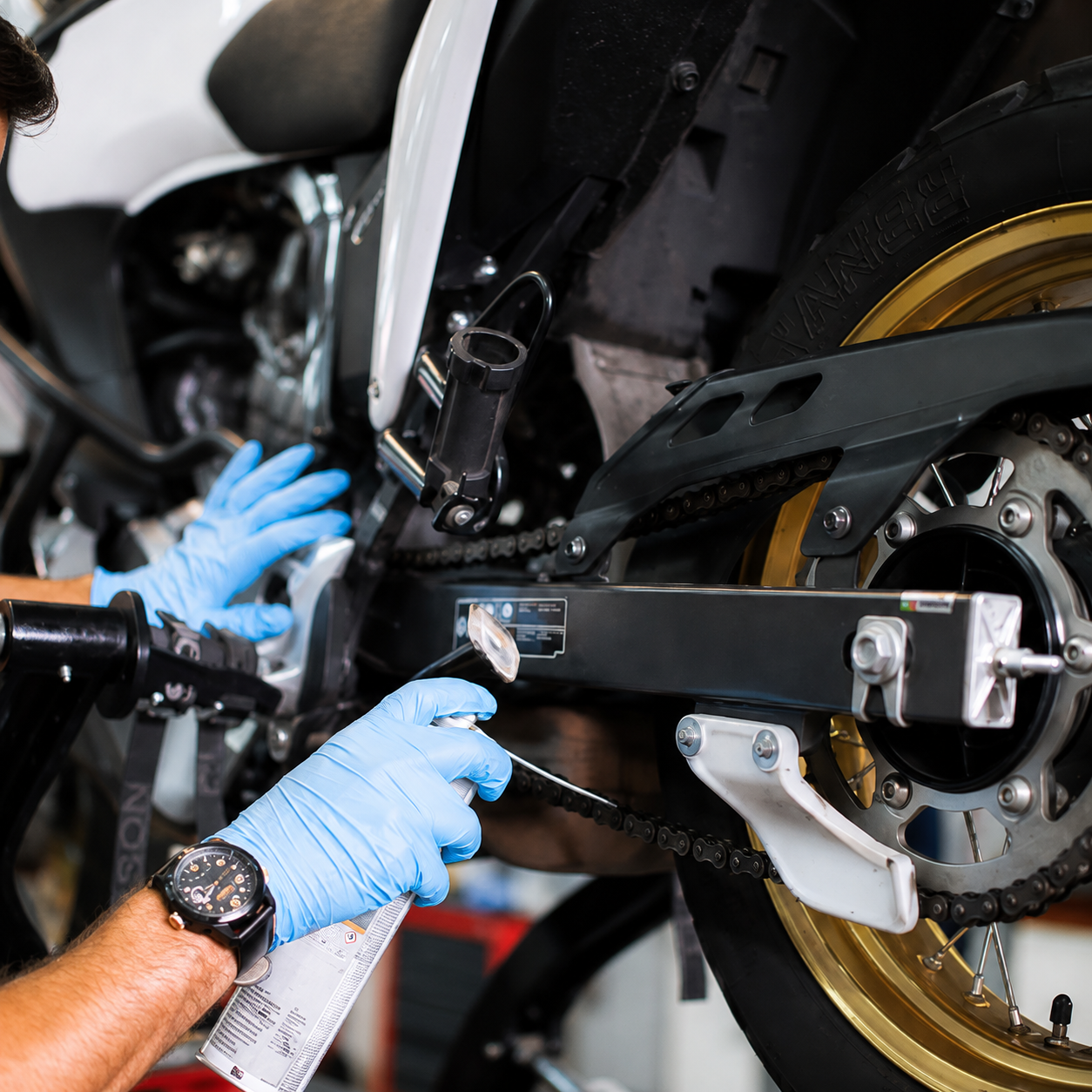 Persoon met blauwe handschoenen voert onderhoud uit aan een motorfiets, gericht op de ketting en het trappergebied.