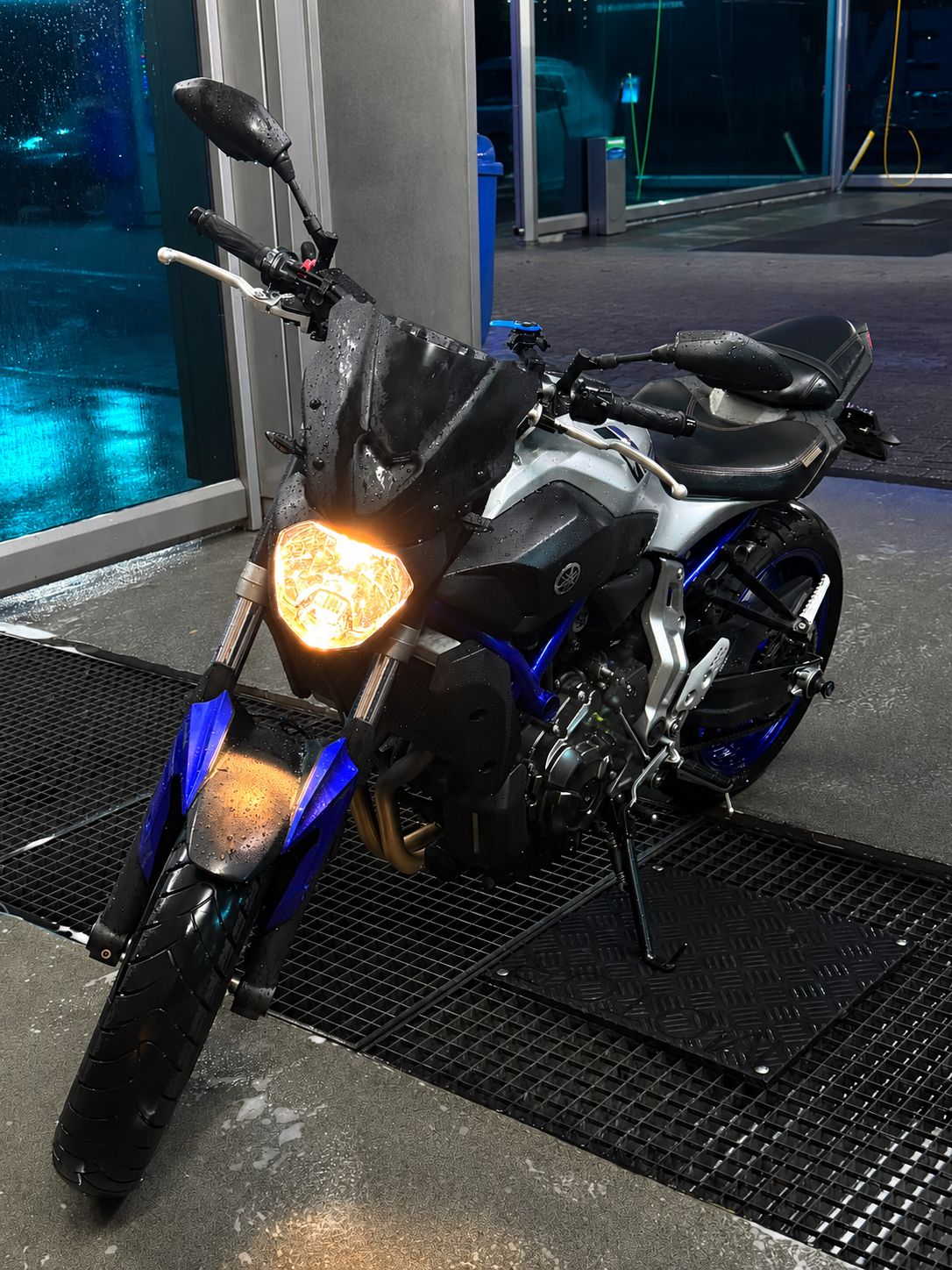 Een sportmotorfiets met een zwart windscherm en blauwe accenten, nat door regen, geparkeerd binnen bij een deur met waterdruppels op de koplamp en enkele reflecties op het lichaam, ’s avonds of ’s nachts.