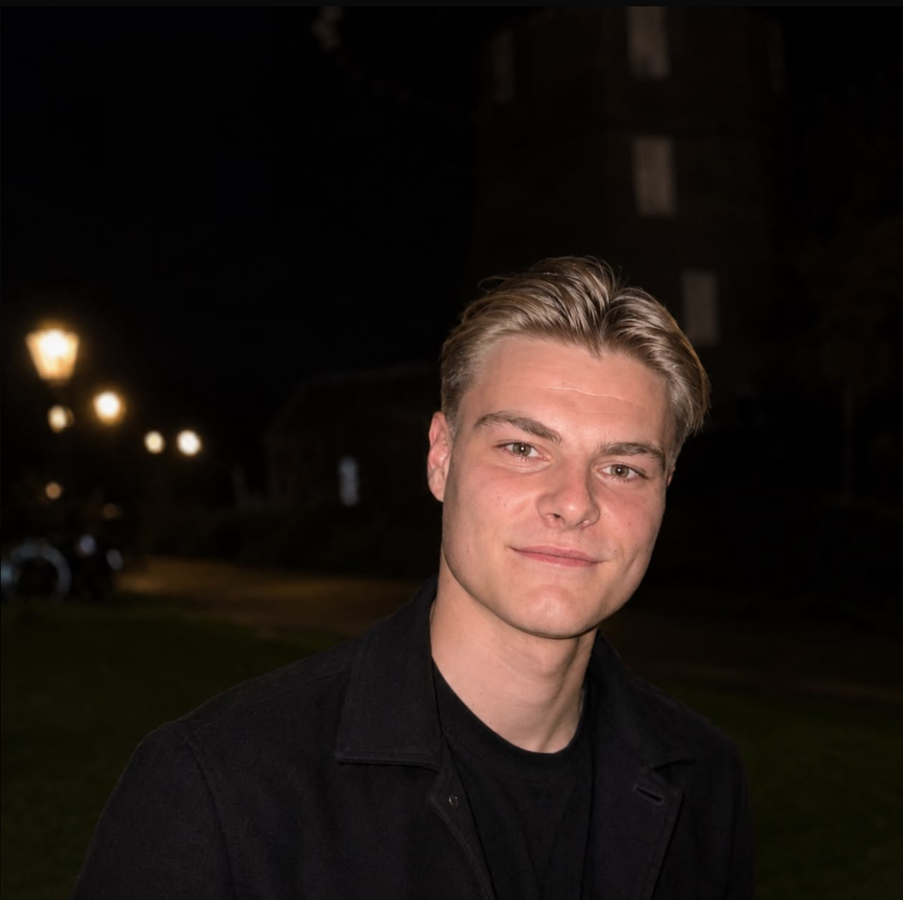 Portret van een jonge man met blond haar, in een zwarte jas, 's avonds buiten.
