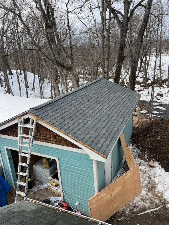 View of a small house under construction with teal siding, a newly installed gray shingle roof, an open garage door with building materials inside, a ladder leaning against the house, and a large plywood sheet on the ground. Surrounding trees and sno