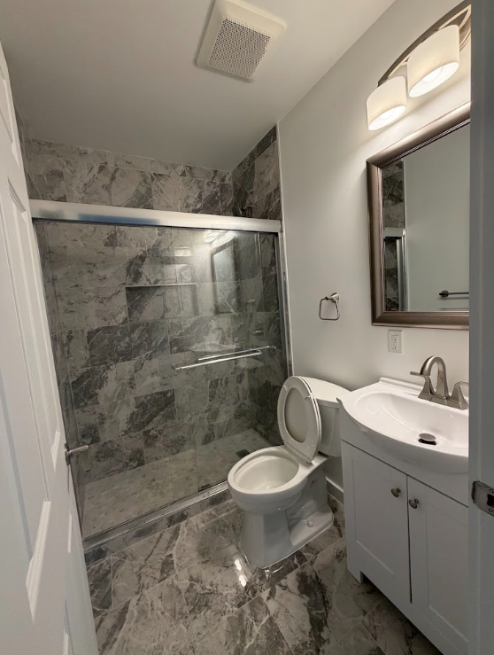 Bathroom with a marble floor and shower, toilet next to a small white sink with a cabinet, mirror, towel holder, and light fixture above.