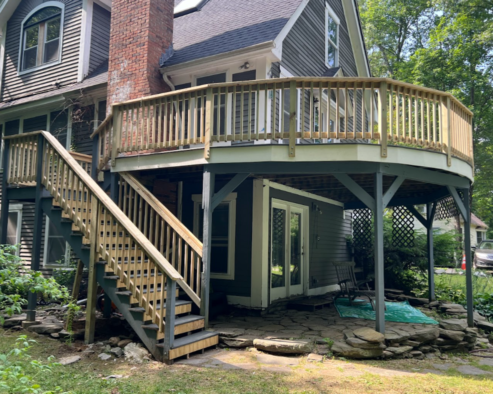 Backyard view of a multi-story house with a new wooden deck and stairs. The house has gray siding, a brick chimney, and white trimming. The ground level has a small outdoor sitting area with a rocking chair, a teal outdoor rug, and a sliding glass do