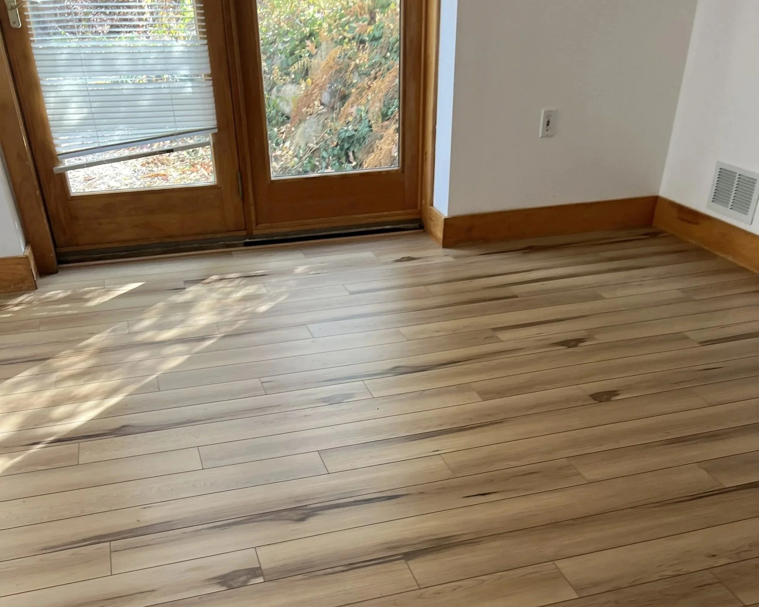Room with wooden floor and a glass door with a white blind, showing leaves outside, and part of a white wall with a wall vent and power outlet.
