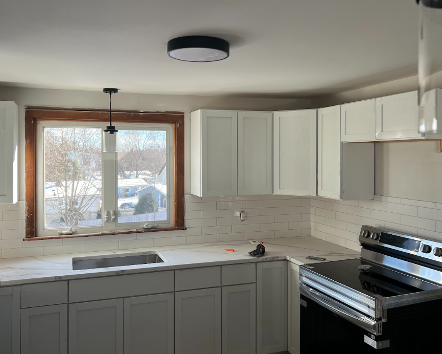 Kitchen with white cabinets, marble countertop, white subway tile backsplash, window over the sink, stove, and partially installed range hood.