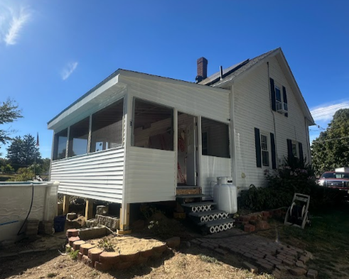 White two-story house with a screened porch, steps leading up to the porch, and a propane tank nearby, set in a yard with brick and stone pavers under a blue sky.