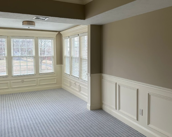 Empty living room with large windows, white wainscot paneling, beige walls, and striped carpet.