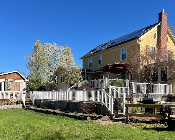 A yellow two-story house with solar panels on the roof, a brick chimney, and a spacious multi-level deck with white railing. The house is surrounded by trees and a vibrant green lawn, under a clear blue sky.