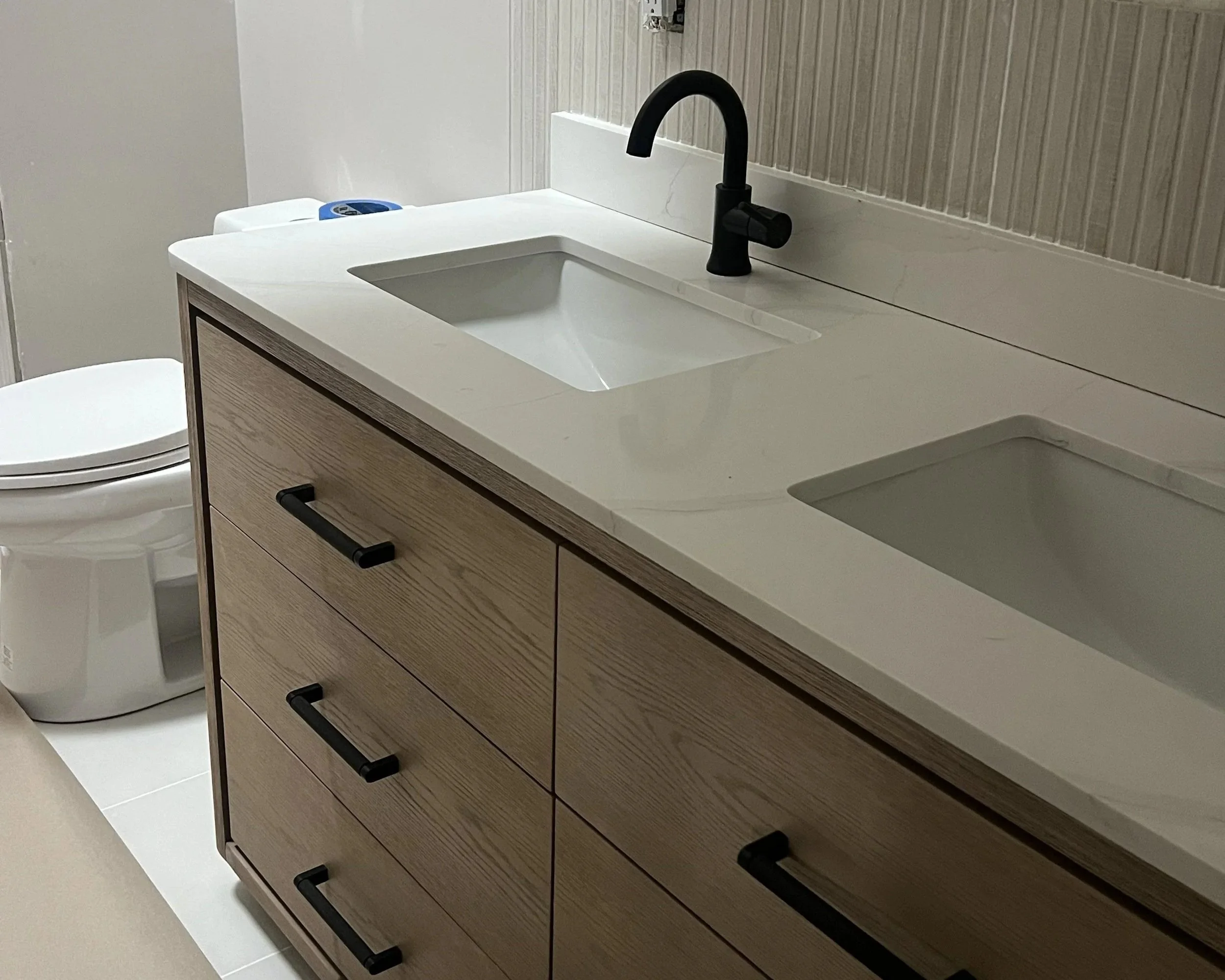 A bathroom vanity with two sinks, a black faucet, and four drawers with black handles. A white toilet is partially visible on the left side of the image. The wall behind the vanity is decorated with vertical beige and white striped wallpaper.