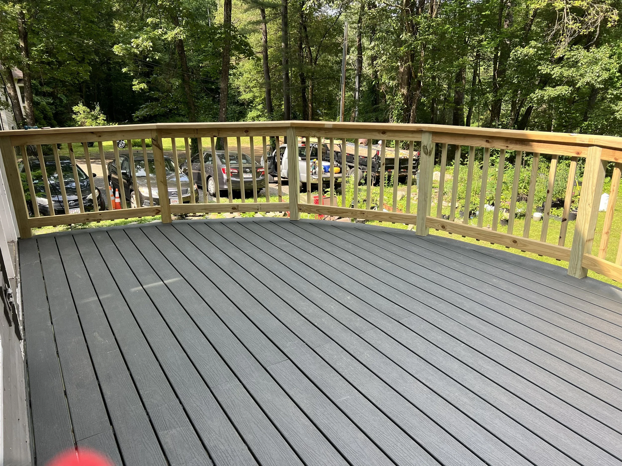 View of a wooden deck railing overlooking a parking lot with cars, surrounded by lush green trees.