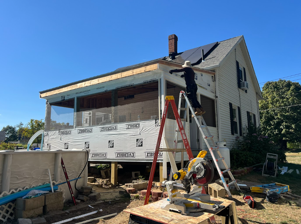 A person working on the roof of a house under construction, standing on a ladder, with construction tools and materials around.
