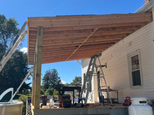 Construction site of a house porch with wooden beams and roofing, ladder, construction tools, and materials on a deck, white house siding, and a clear blue sky.