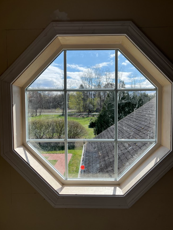 View through a multi-pane octagonal window showing a sunny outdoor scene with trees, grass, a brick patio, and part of a neighboring roof.