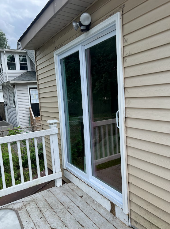 Exterior of a house with a sliding glass door leading to a small porch with white railing, beige vinyl siding, and an outdoor light fixture on the wall.
