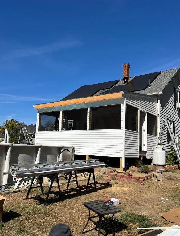 House under renovation with a new porch addition, construction tools, and equipment in the yard, blue sky overhead.
