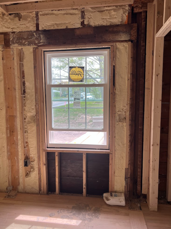 Interior of a house under construction with a window installed in a wall with exposed insulation and framing. The window has a sticker on it, and there is a roll of insulation or protective material on the floor near the wall.