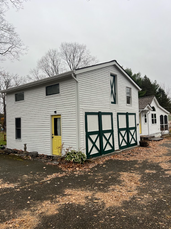 White barn-style garage with green double doors, a yellow door, and black asphalt driveway, situated in a residential area with trees in the background.