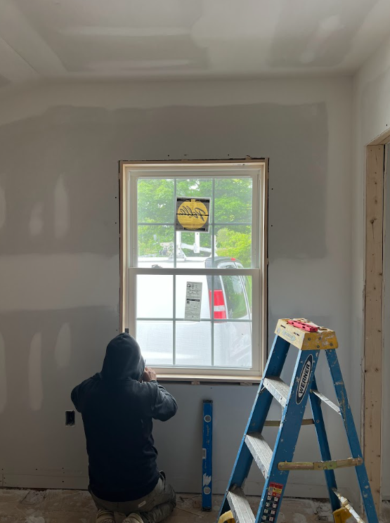 A person working on a window installation inside a room under construction, with a blue step ladder nearby and a vehicle visible outside through the window.