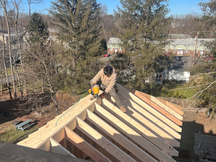 A worker installing roof rafters on a house, sitting on the roof with a power drill, surrounded by trees and residential buildings.