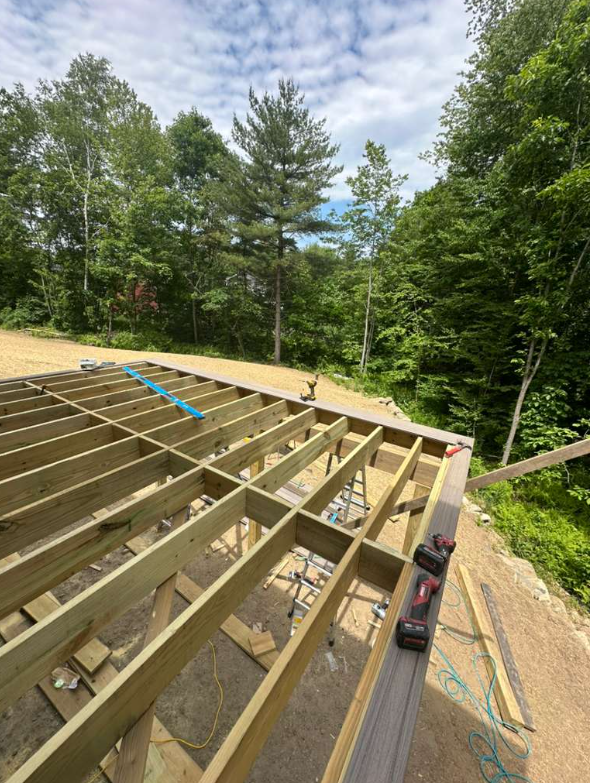 Construction of a wooden deck with framing beams in a wooded area, partial floor structure visible, tools and equipment present.