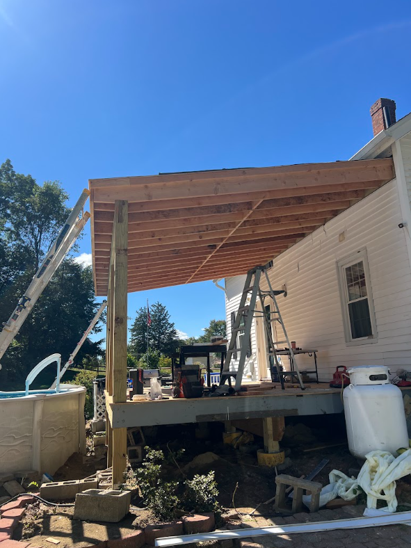 Construction site of a house's porch with wooden support beams and a roof, ladder, tools, and construction materials around.