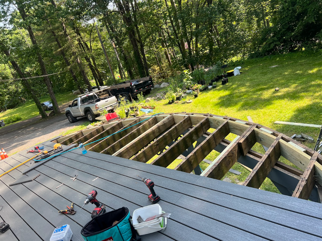 Construction site on a deck with tools, drill, and supplies, with a partially built wooden section and a green backyard with trees and parked vehicles.