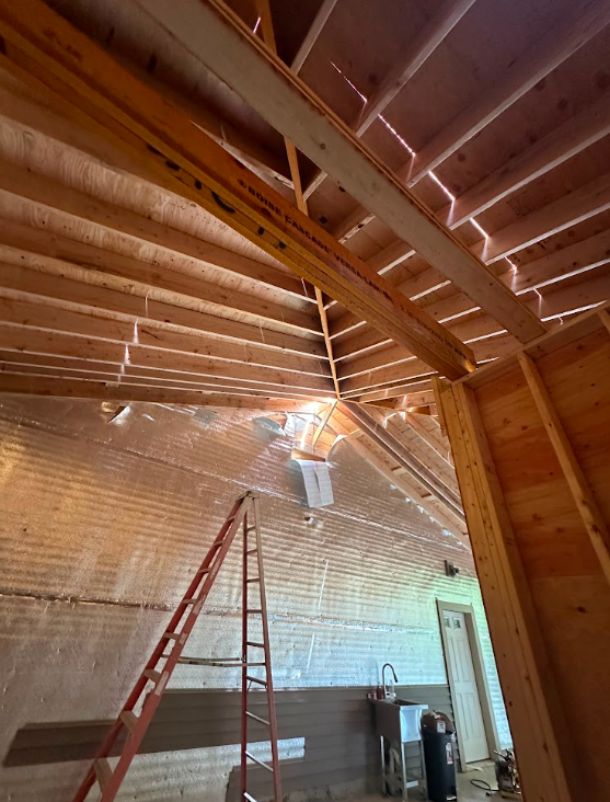 Interior construction of a wooden building, showing exposed roof rafters, a ladder, and a wall with insulation and a vent. A door and a utility sink are also visible.