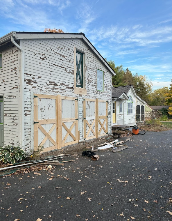 A house undergoing renovation with part of the exterior siding removed and new wooden panels installed. There are trees and a partly cloudy sky in the background, and construction debris on the asphalt driveway.