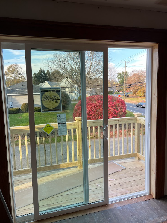 View of a neighborhood through a sliding glass door with a wooden deck outside. There are houses, trees, a sidewalk, a street with cars, and a large red bush in the background.