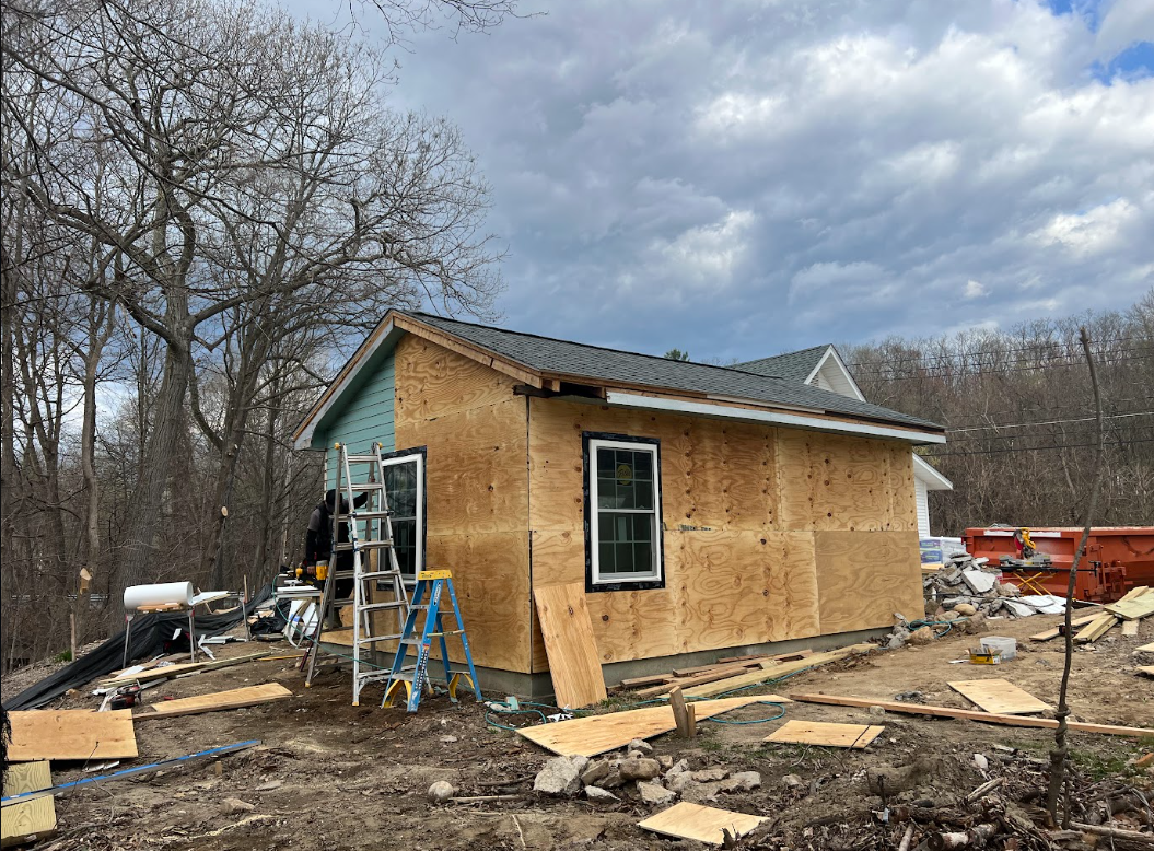 A house under construction with plywood walls, a window, and a partially finished roof, surrounded by construction tools and debris, with leafless trees and a cloudy sky in the background.