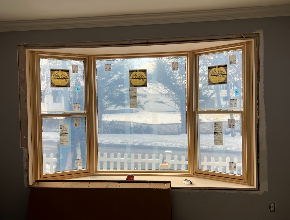 Newly installed bay window with sticker labels, overlooking a snowy yard with trees and a white picket fence.