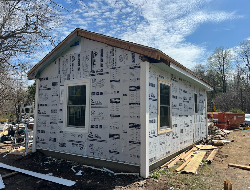 Construction site of a house with exterior walls covered in protective sheathing, two windows installed, and a ladder leaning against the side.