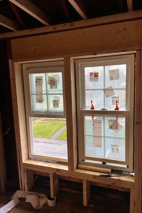 Two new double-hung windows installed in a wood frame wall, with stickers and tags attached to the glass, and two red spray bottles on the window sill, in a house under construction.