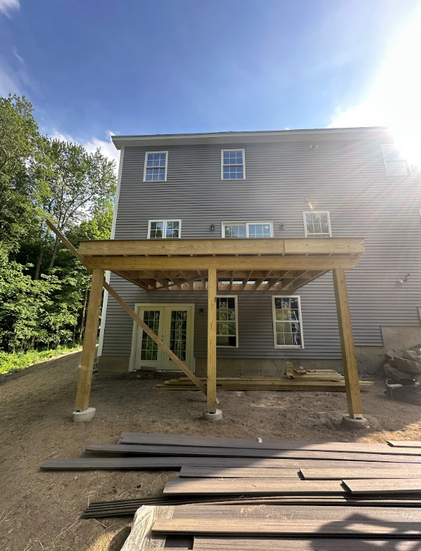 Construction of a wooden deck on the back of a gray three-story house with multiple windows, surrounded by trees on a sunny day.