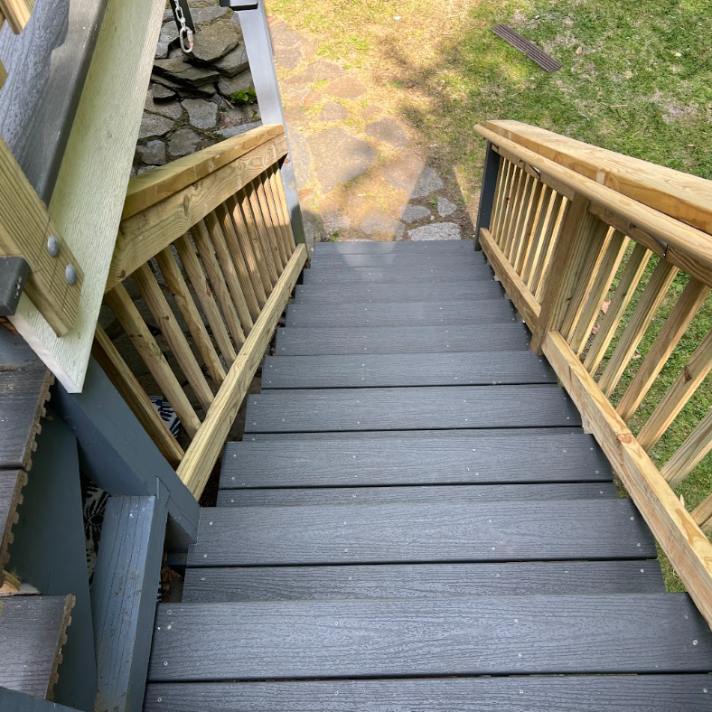 Wooden outdoor staircase with black painted steps and wooden railings leading down to a stone and grass yard.