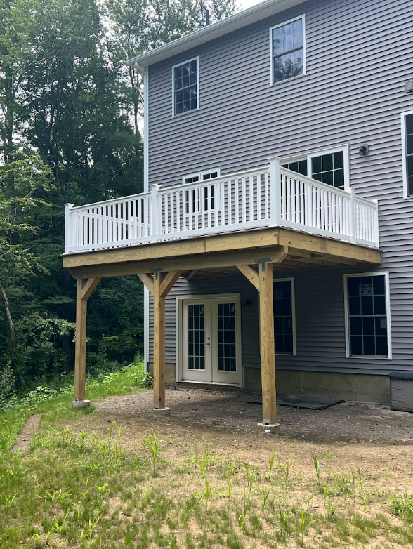 New wooden deck construction attached to the back of a gray house with white siding. The deck has a white railing and is supported by four wooden posts. The house has multiple windows and a set of double glass doors leading to the ground level. The y