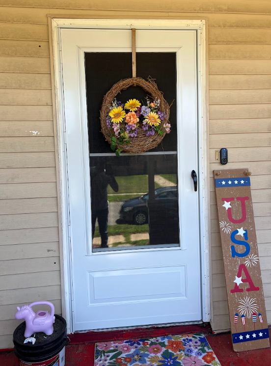 Front door with a wreath of colorful flowers, a colorful welcome mat, a purple watering can, and an 'USA' wooden sign with patriotic decorations.