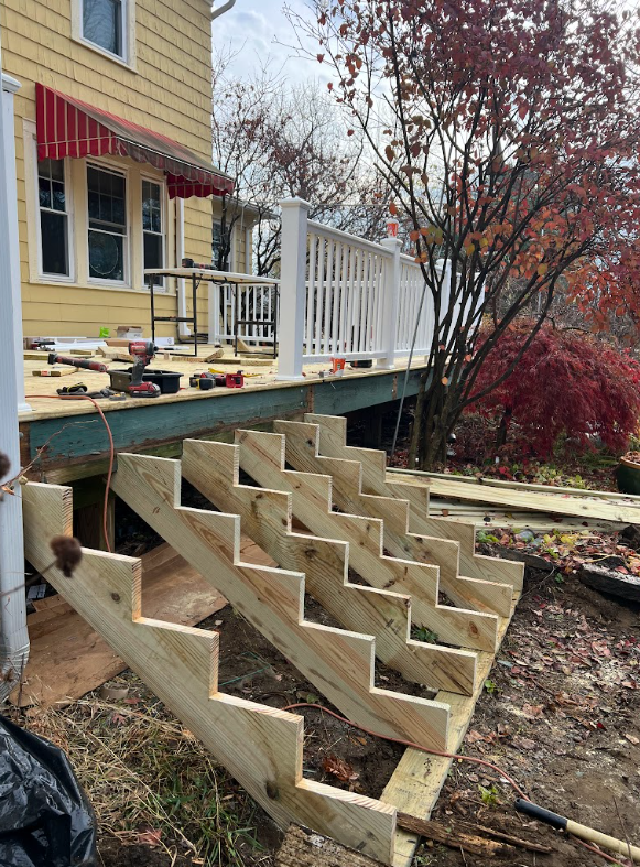 Wooden staircase framing under construction outside a house. The house has a porch with a red awning, a white railing, and yellow siding. There are tools on the porch and trees with fall foliage in the background.