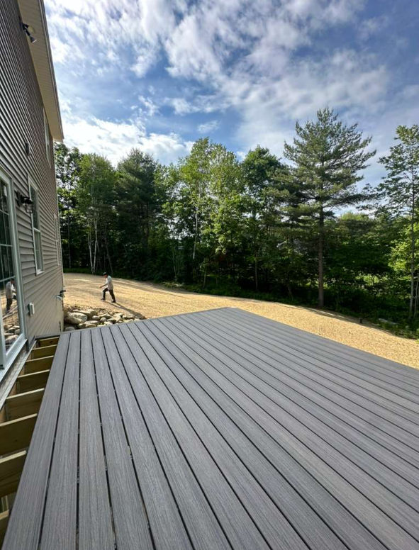 A backyard with a newly constructed gray deck, a gravel pathway, and a wooded area with tall trees under a partly cloudy sky. Two people are seen walking along the gravel path.