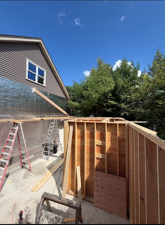 Construction site with wooden framing for a new structure, ladders, and building tools, next to a house and trees under a blue sky.