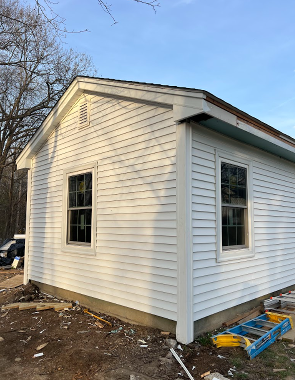 A white house with vinyl siding, two double-hung windows, and a gable roof under a clear blue sky. Construction materials are on the ground around it.