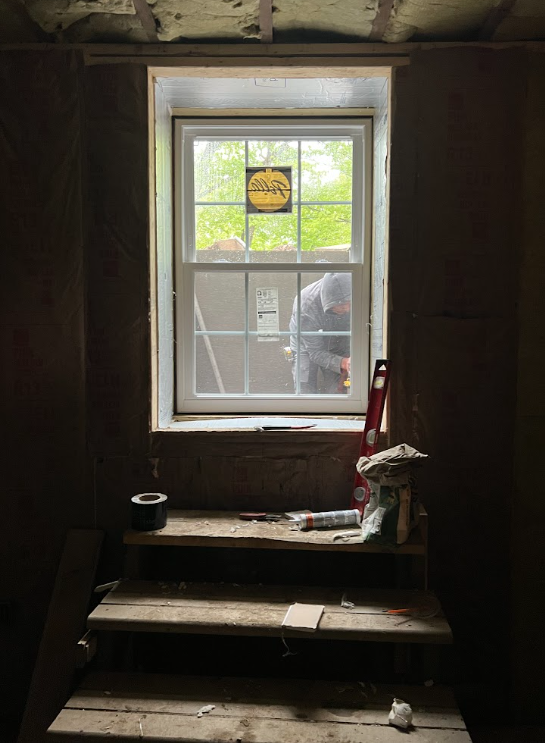 View through a new window in a construction site, showing a worker outside. The window has a yellow sticker at the top center, and the room has unfinished interior with tools and materials on wooden steps.