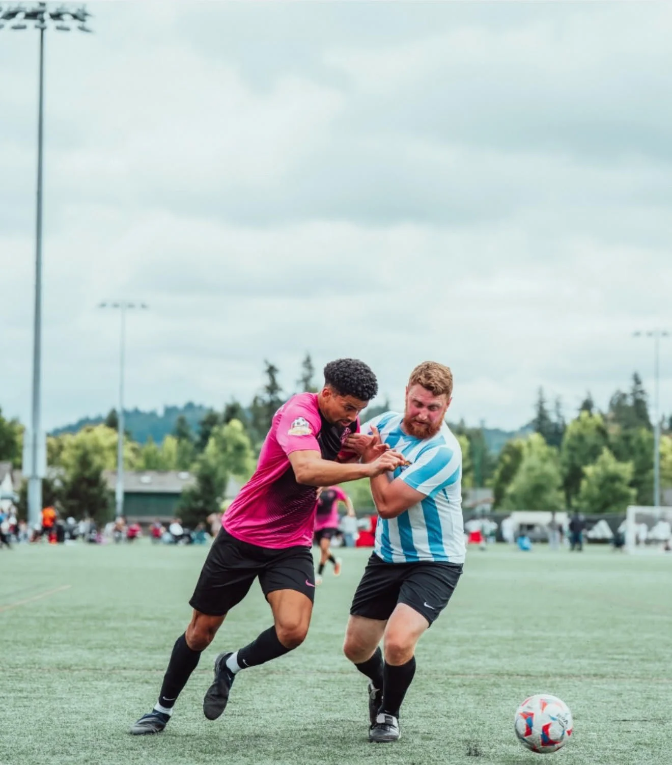 Two soccer players competing for the ball during a match on a grassy field, with spectators and trees in the background under a cloudy sky.
