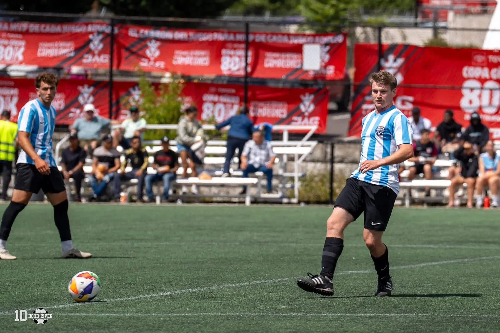 Two soccer players with light skin and dark hair, wearing blue and white striped jerseys and black shorts, on a green artificial turf field during a game, with spectators sitting on white bleachers behind them and red advertising banners in the background.