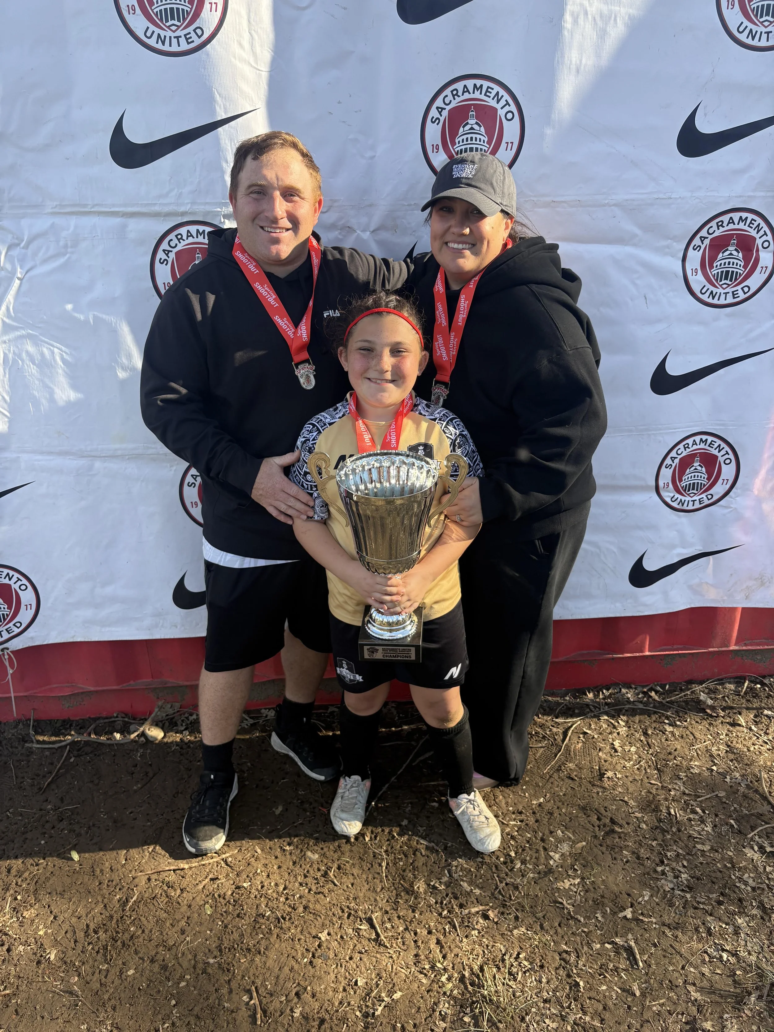 Three people standing together, holding a trophy and medals, in front of a backdrop with Sacramento United logos, celebrating a sports achievement.